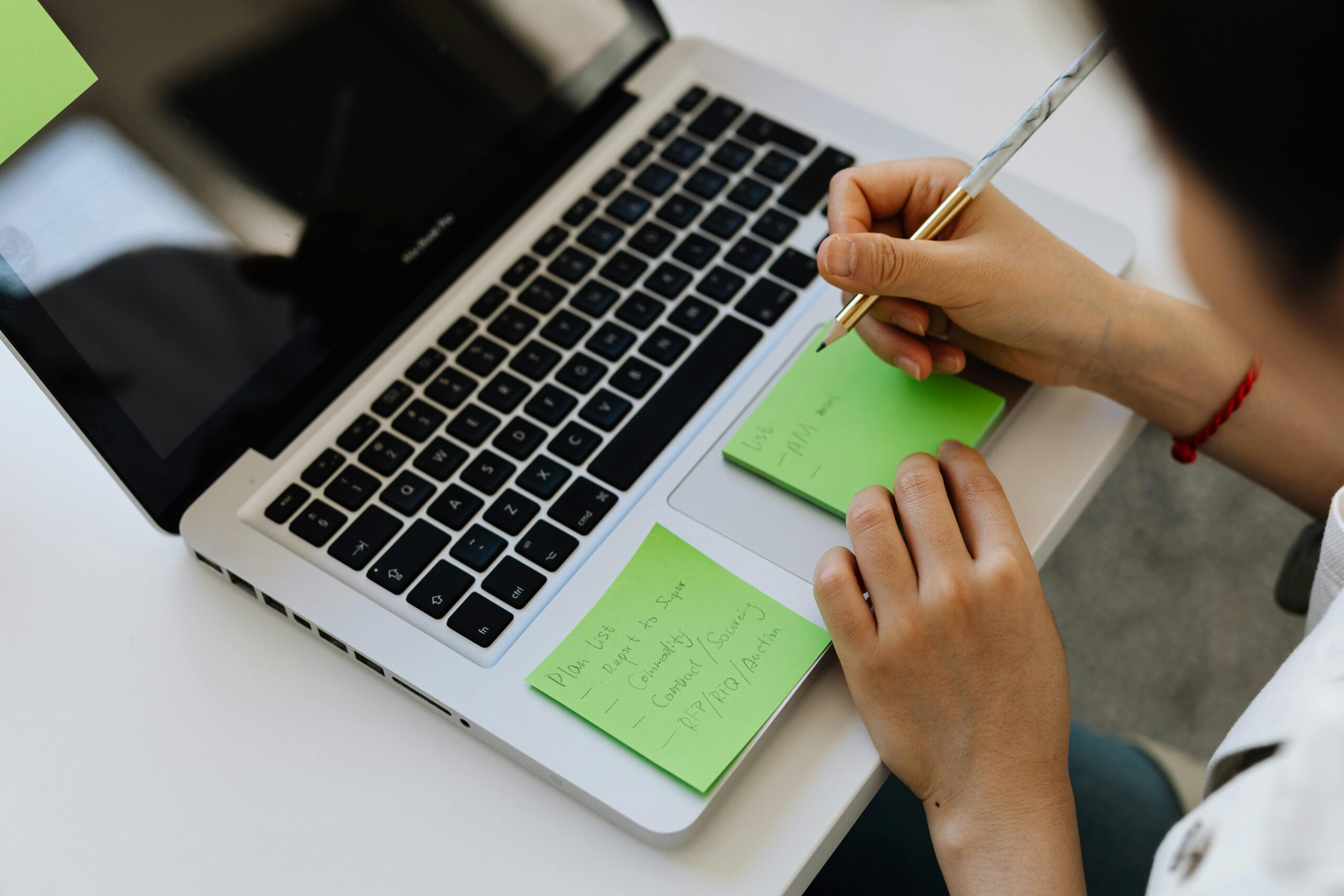 An individual writes notes on green sticky notes placed on a laptop in a work setting.